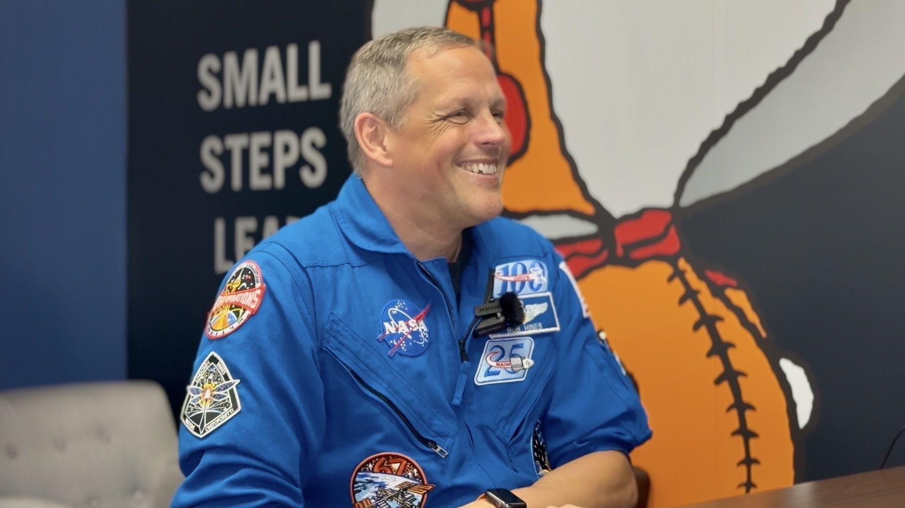 A man in a blue jumpsuit sits smiling in front of a wall with a giant snoopy astronaut painted on.