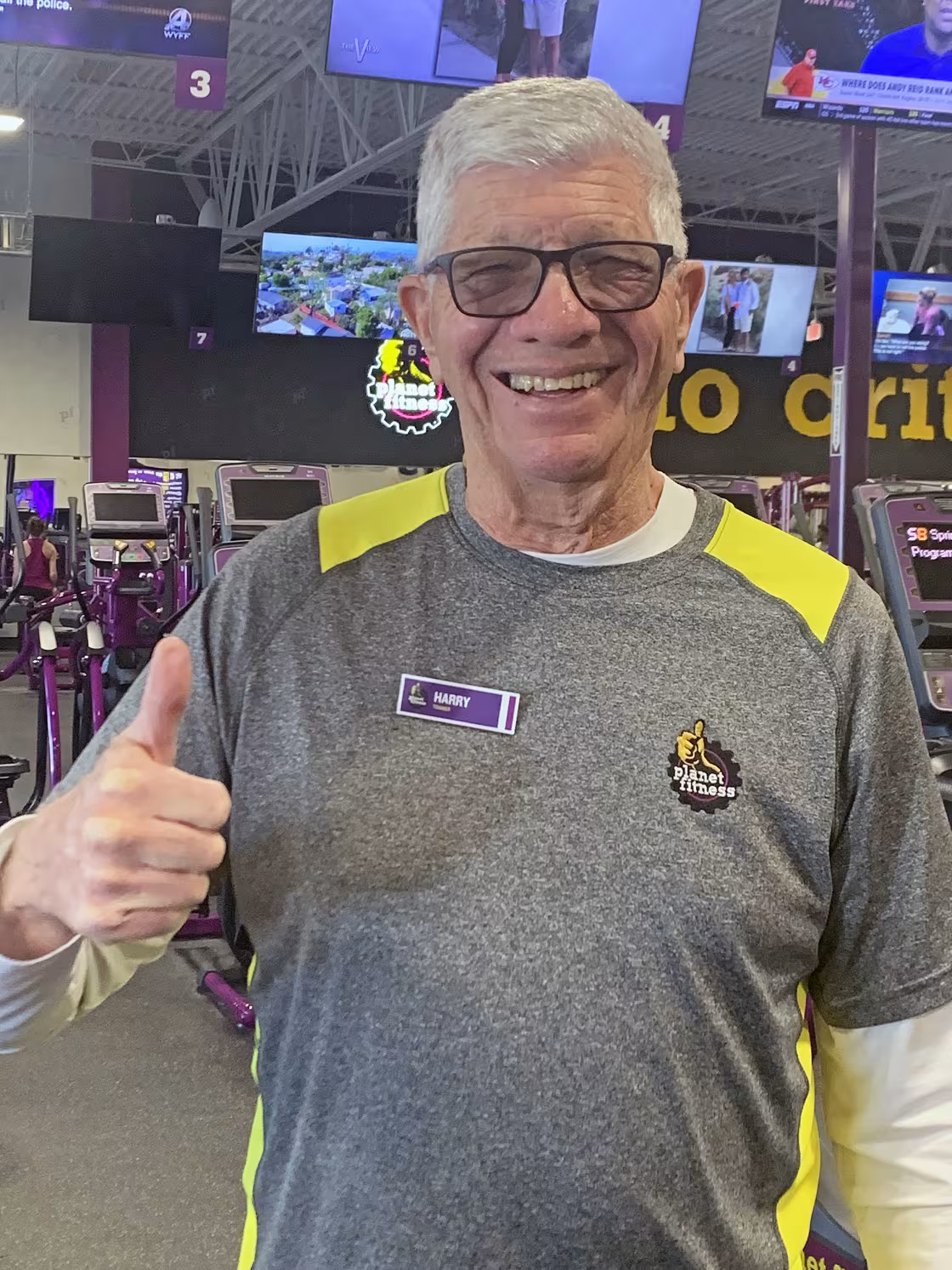 Man wearing Planet Fitness t-shirt with nametag that reads Harry stands in a gym and gives a thumbs up