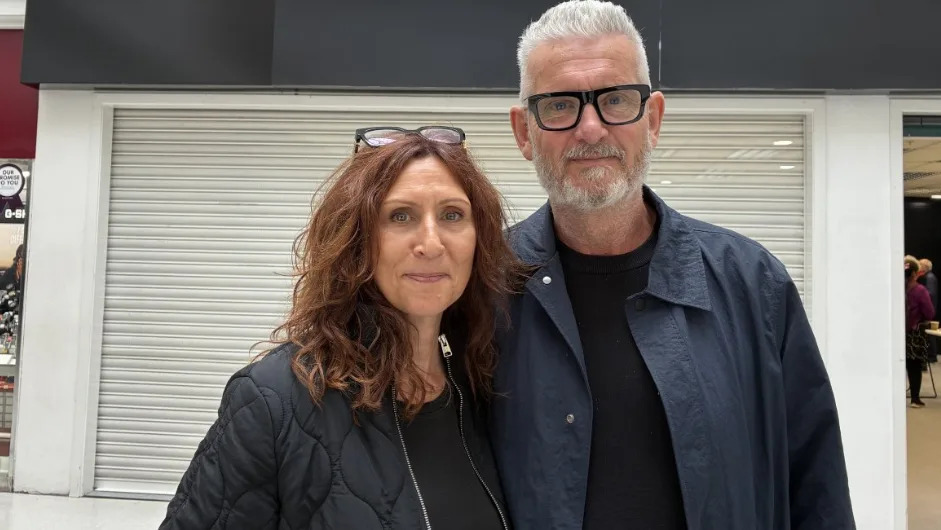 A woman with long brown curly hair and glasses on her head stood next to a man with silver hair and black glasses. They are both wearing black tops and navy jackets.