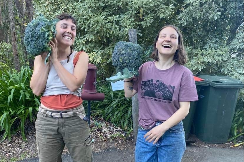 Two laughing women hold up very large broccolis.