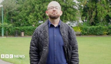 A bald and beared bespectacled man in his thirties, wearing a blue shirt and trousers and a dark jacket, stands in a park with his hands in his pockets.