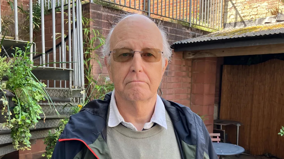 The image shows a grey haired man, wearing tinted glasses and a blue and green coat, standing in a courtyard.