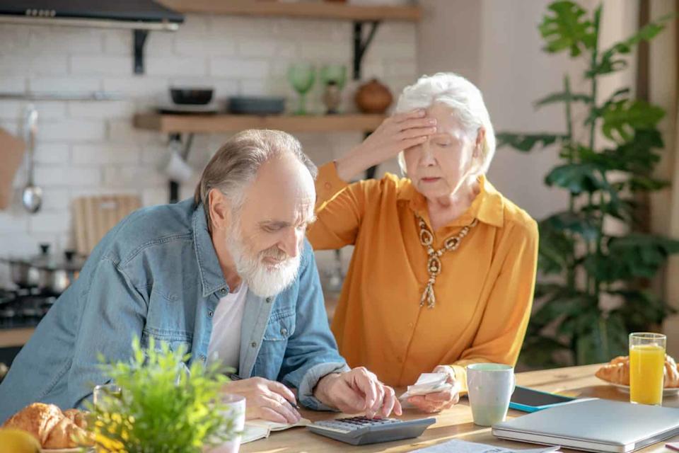 A senior couple in a kitchen. The man with a white beard is seated, writing in a notebook and using a calculator. The woman, with white hair, stands beside him, her hand pressed to her forehead with a distressed expression. On the wooden table are papers, a coffee mug, a laptop, croissants, and orange juice.