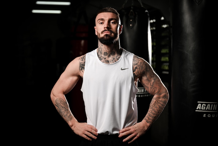 Boxer Lewis Crocker ikn a white singlet, in a gym, posing with hands on hips