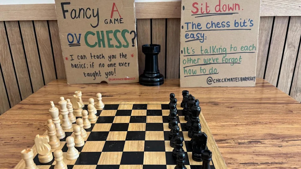 A chess board on a wooden table with two homemade signs propped up inviting people to play.