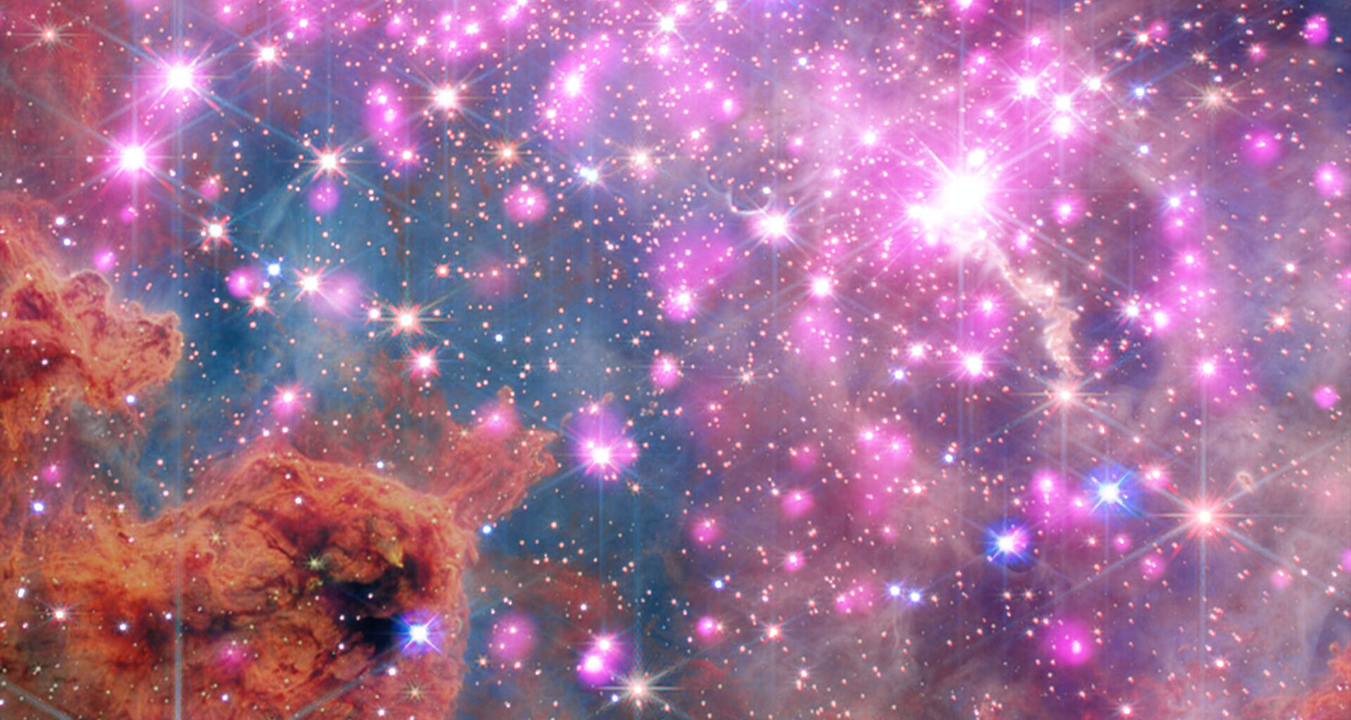 Scores of gleaming stars ringed in neon pink stretch across the frame, highlighting a cluster where stars are between one and three million years old. Brick-orange dust clouds along the bottom edge illustrate the raw materials of this active stellar nursery.