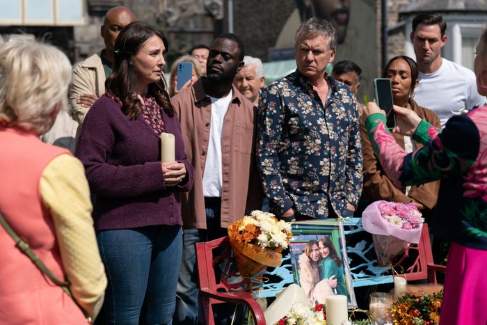 566590,TITLE:Eastenders Linda films a vigil for Honey, where she stands over a bench with some flowers and a picture of her and Bea in a scene from EastEnders