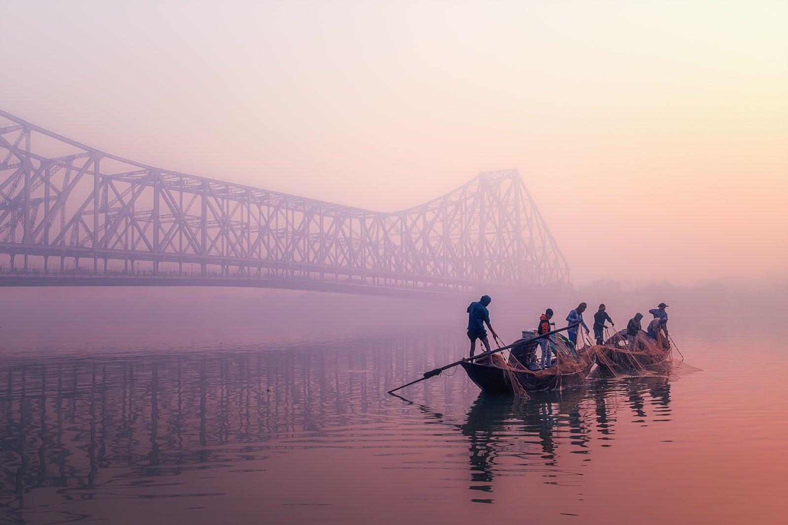 A group of people row a wooden boat on a calm river at sunrise, with the Howrah Bridge visible through the mist in the background. The scene is peaceful, with soft pastel colors filling the sky.