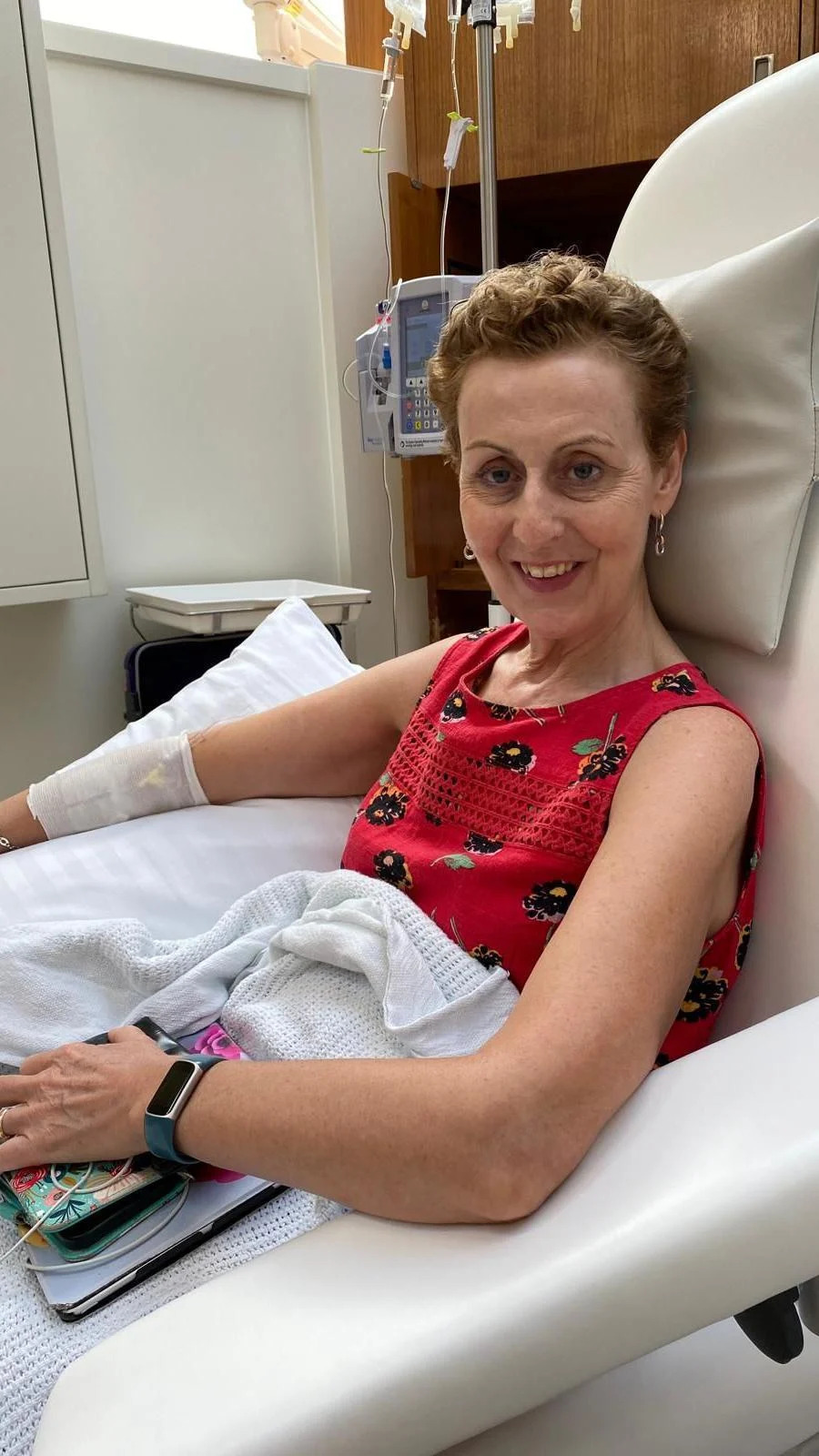 The image shows a person sitting in a reclining medical chair in a clinical setting. The person is wearing a sleeveless red top with a patterned design and has a blanket draped over their lap. An intravenous line is inserted into the person’s arm, secured with a bandage, and connected to medical equipment mounted behind the chair. The person’s arm rests on the chair’s armrest, and a small bag or personal items are placed nearby. The room has clean white and wooden surfaces, with medical devices and monitors visible in the background.
