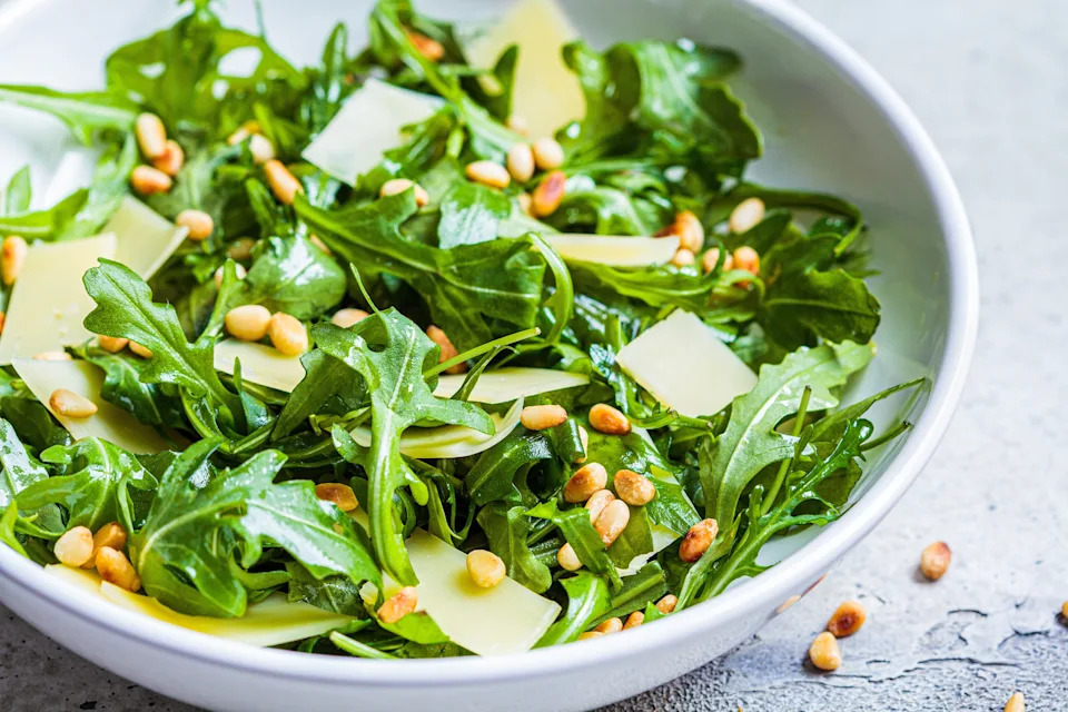 A fresh salad in a bowl with arugula, shaved parmesan, and pine nuts on a textured surface