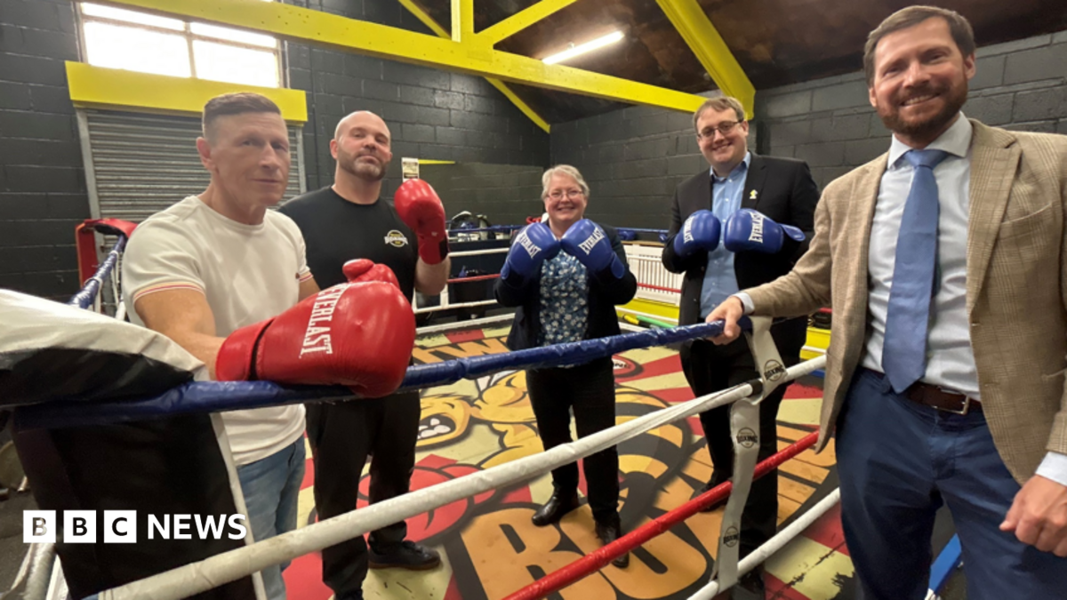 Three men and a woman standing in a boxing ring, and one man standing just outside it. The ones inside are wearing boxing gloves and they are all smiling.