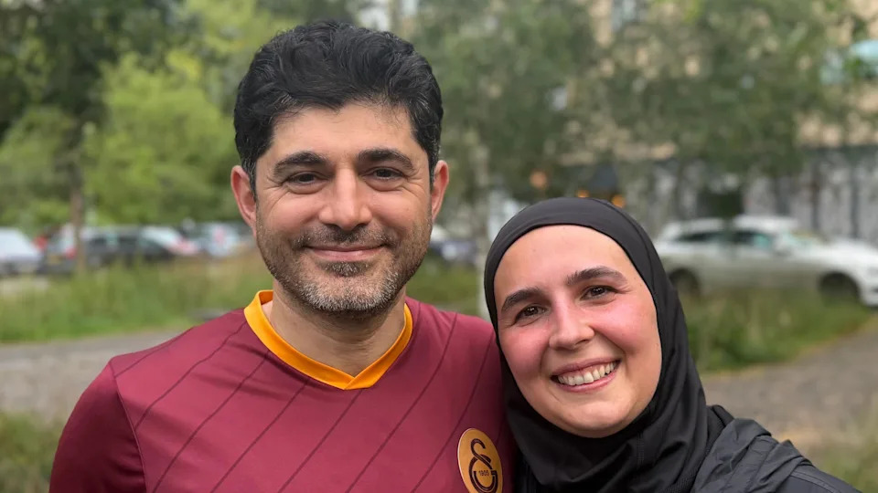 A man and a woman smiling straight at the camera. There are trees and parked cars behind them. The man is wearing a burgundy and orange T-shirt and the woman is wearing a black hijab.