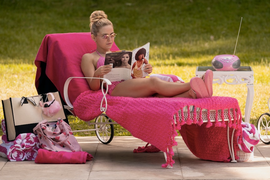 Lexi Minetree as Elle sitting on a pink shawl outside reading a magazine surrounded by 90s memorabilia