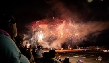 Nighttime stadium spectators watch a fireworks display above the track, with bursts of red, gold, and white lights.