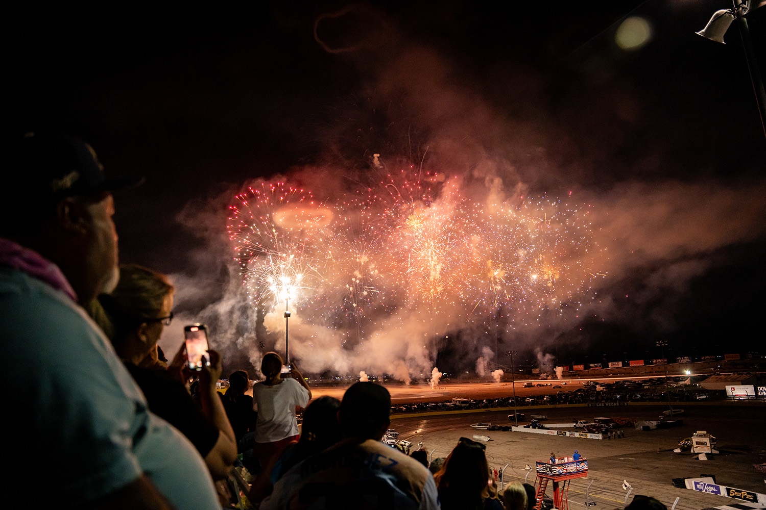 Nighttime stadium spectators watch a fireworks display above the track, with bursts of red, gold, and white lights.
