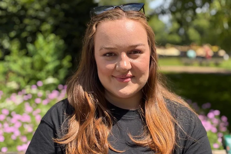 A woman in her late twenties with long hair, wearing a black top, smiling at the camera. She's sat in a public park with greenery behind her.