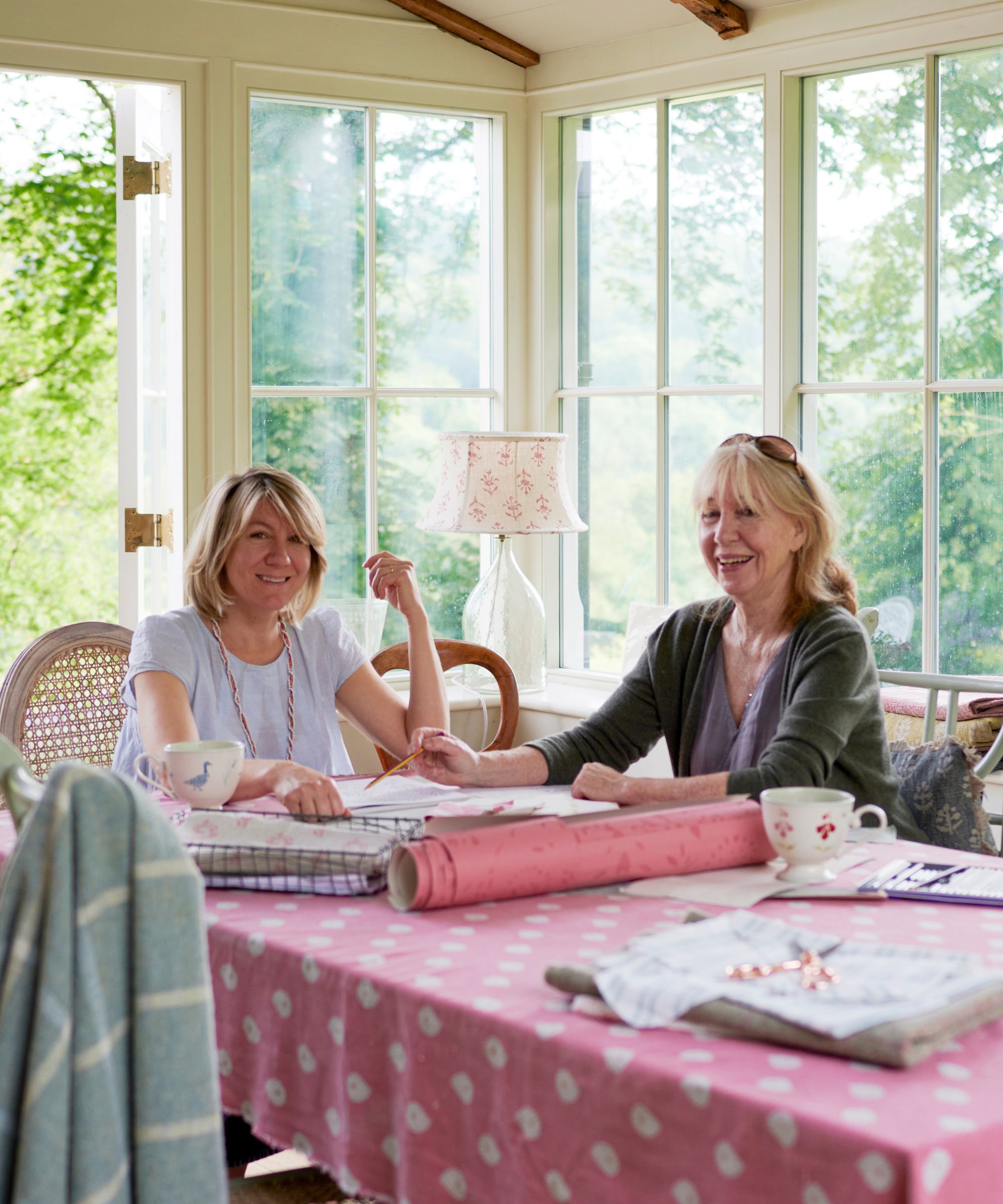 Designers Susie and Anna Watson smiling in a bright sunroom while reviewing design sketches and fabrics at a pink table.