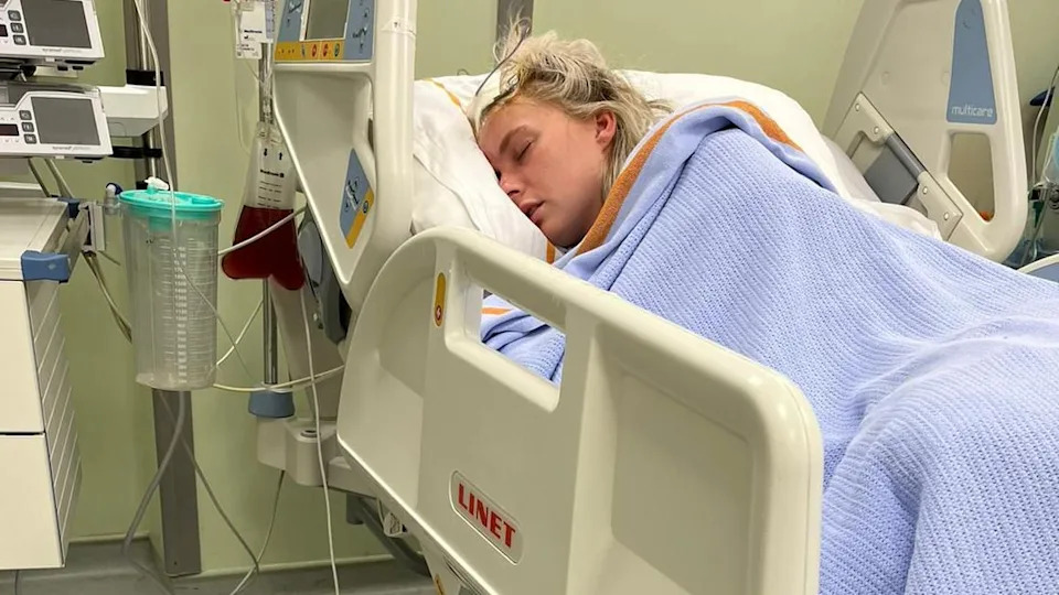 Mererid is in a hospital bed with a blue blanket covering her and a white dressing on her head after surgery.