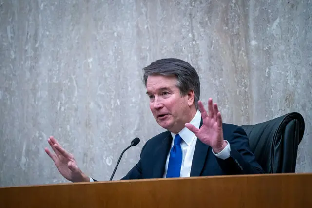 Brett Kavanaugh behind a microphone wearing a blue tie with his hands up