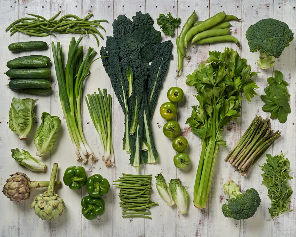 Various green vegetables, including cucumbers, broccoli, kale, lettuce, artichokes, celery, beans, and peppers, arranged on a rustic wooden surface
