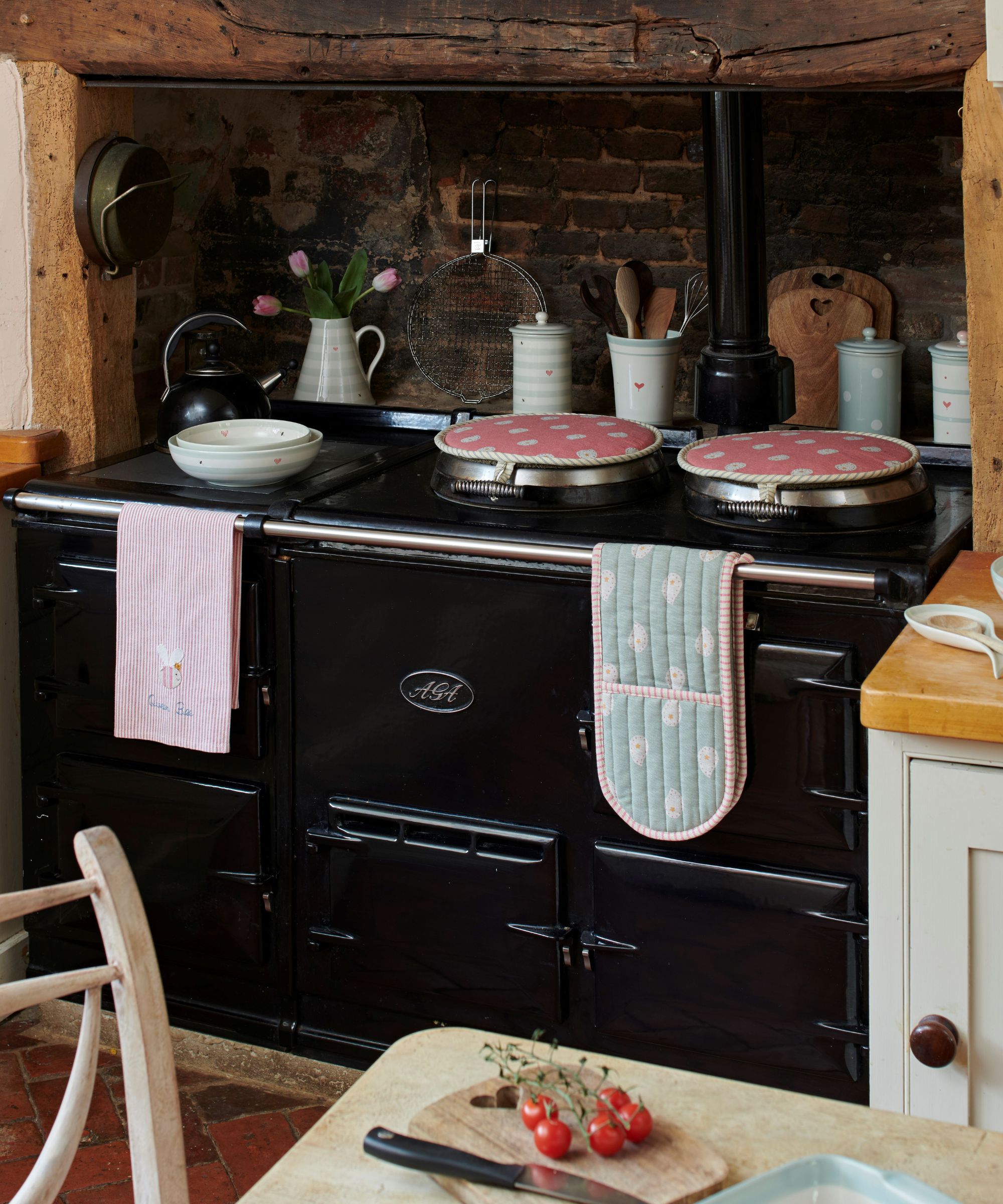 A classic black AGA cooker in a brick alcove, styled with pink and green striped textiles, ceramic jars, and fresh tulips