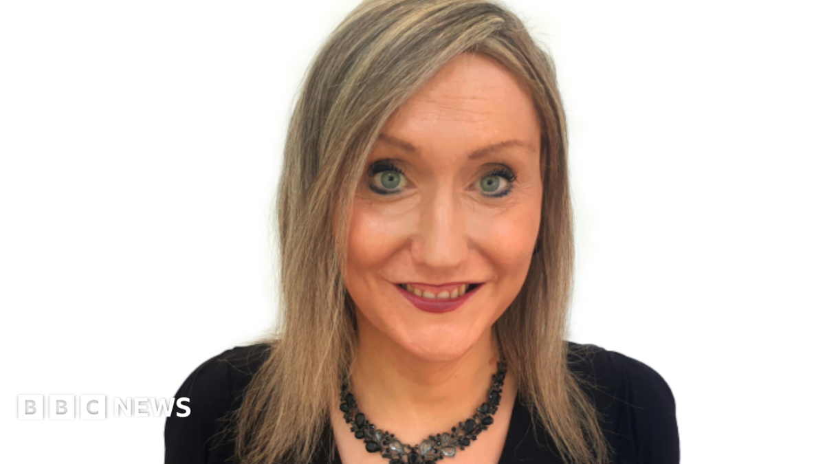 Lisa McNally pictured in front of a white background. She has shoulder length dark blonde hair and is wearing a black necklace and black top.