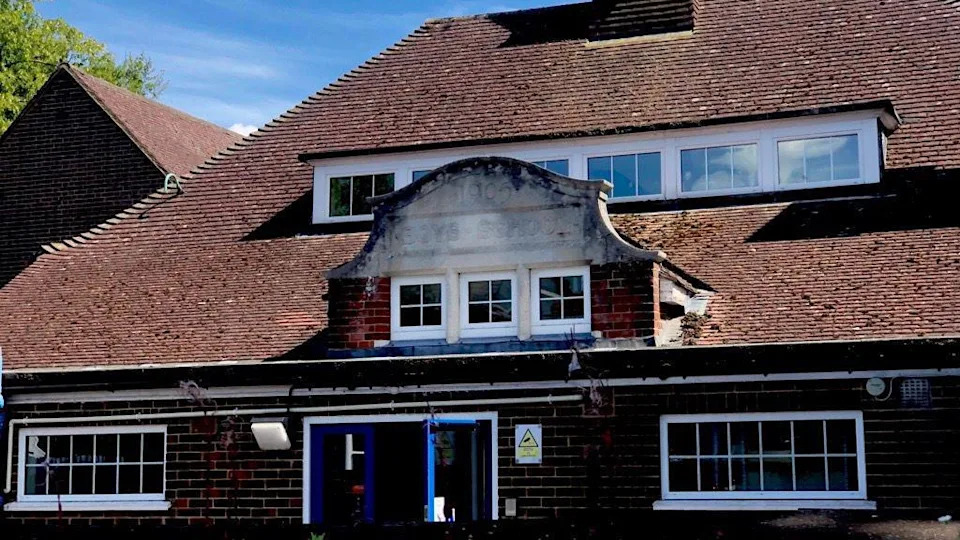 Outside of an infants school in Surrey. A red/brown brick building, which has faded letters in the middle, which say: 1905, BOYS SCHOOL'