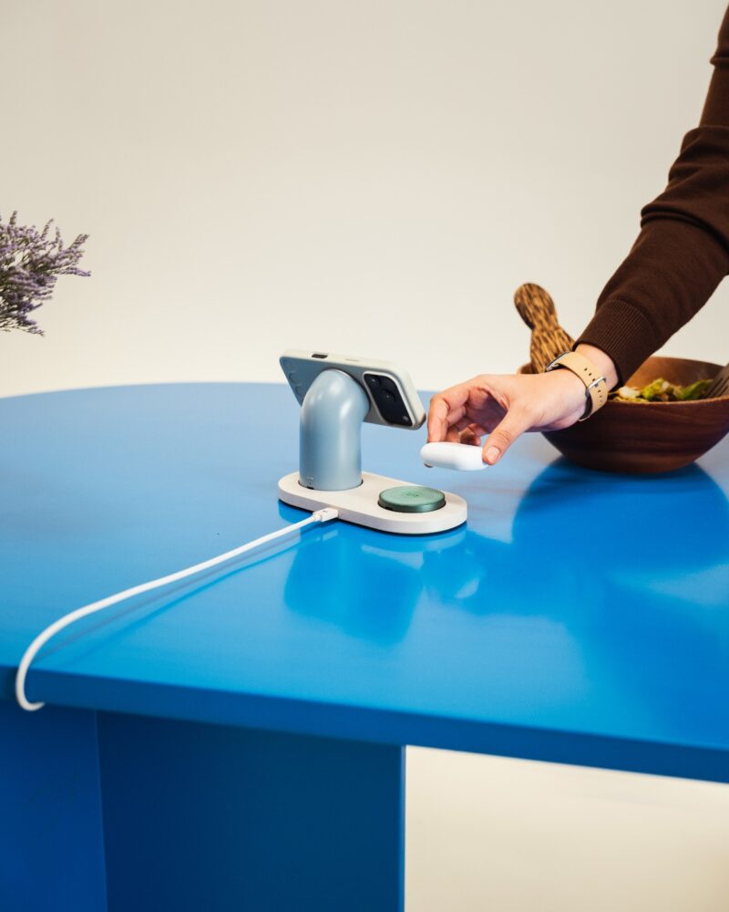 A person’s hand touches a DailyObjects circular device on a blue table, next to a smartphone on a charging stand and a bowl of food.