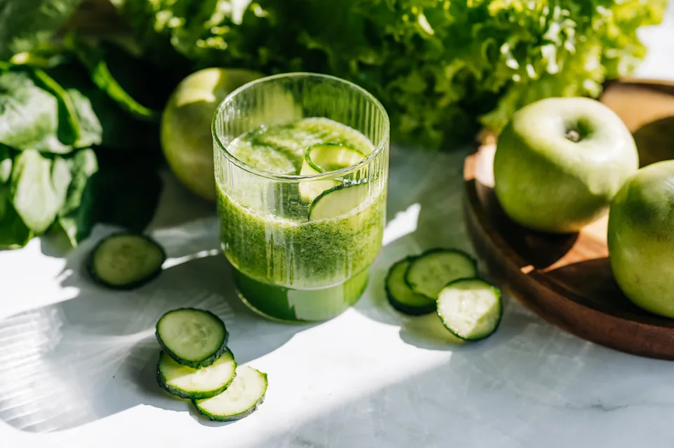 A glass of green juice with cucumber slices on top, surrounded by fresh green apples, cucumbers, and leafy greens on a bright surface