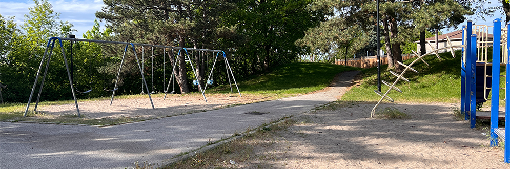 Existing playground at cayuga Park featuring a swing set, climbing structure, and paved path leading up a grass hill, surrounded by mature trees.