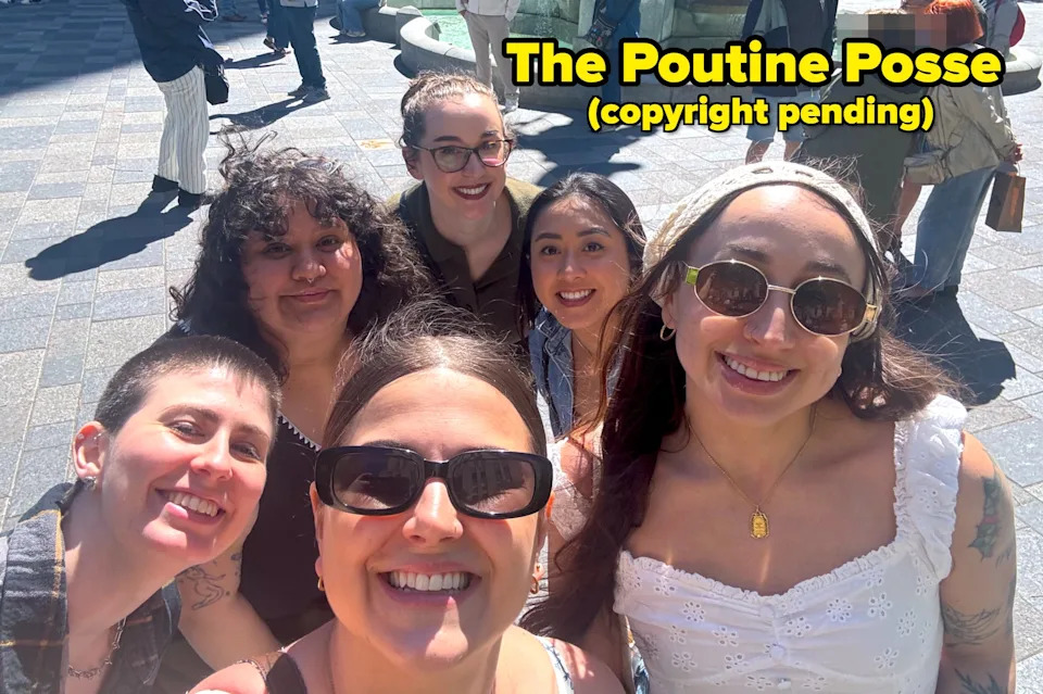 Group of six smiling friends taking a selfie in front of a fountain in an outdoor plaza