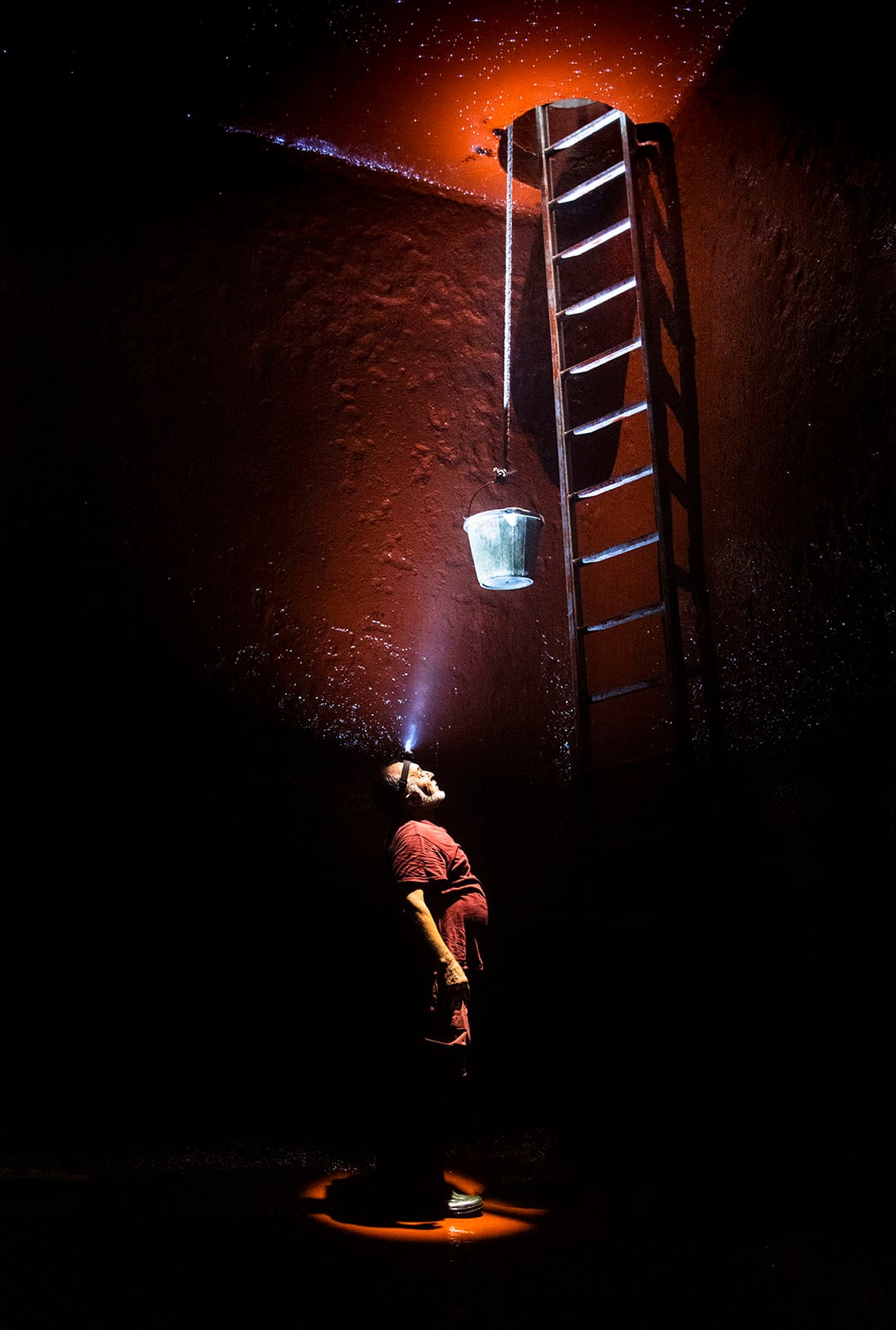A person wearing a headlamp stands under a ladder, looking up at a metal bucket suspended from the ceiling, illuminated by a beam of light in an otherwise dark, reddish-toned space.