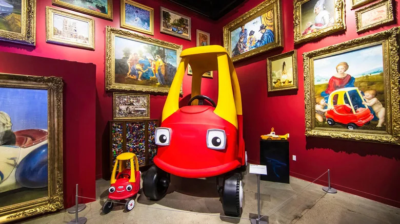 2024-11-15 Los Angeles USA A life-sized version of the iconic Little Tikes Cozy Coupe, displayed alongside a smaller toy version in a museum exhibit, surrounded by classic framed paintings.