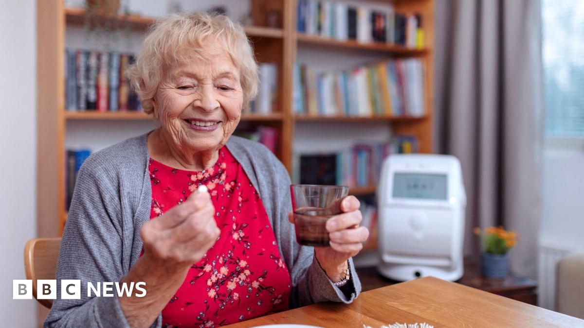 An elderly woman wearing a red floral dress and grey cardigan. She is holding a glass of water and a pill. She is sat at a wooden table. Behind her is a white wall and bookshelf.