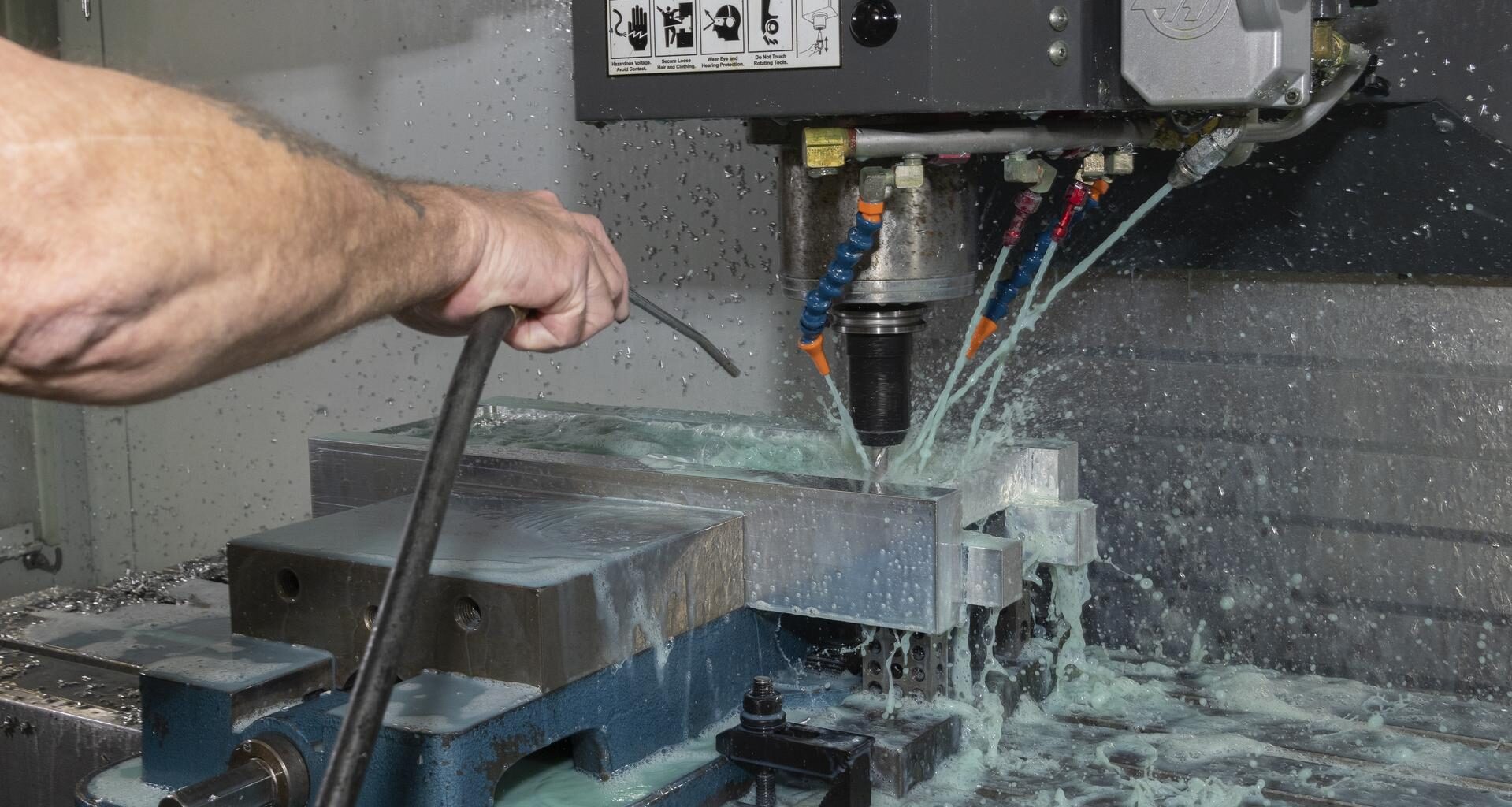 A technician is using a machine to drill pieces of silver metal; there is a light blue liquid coming out of jets next to the drill.