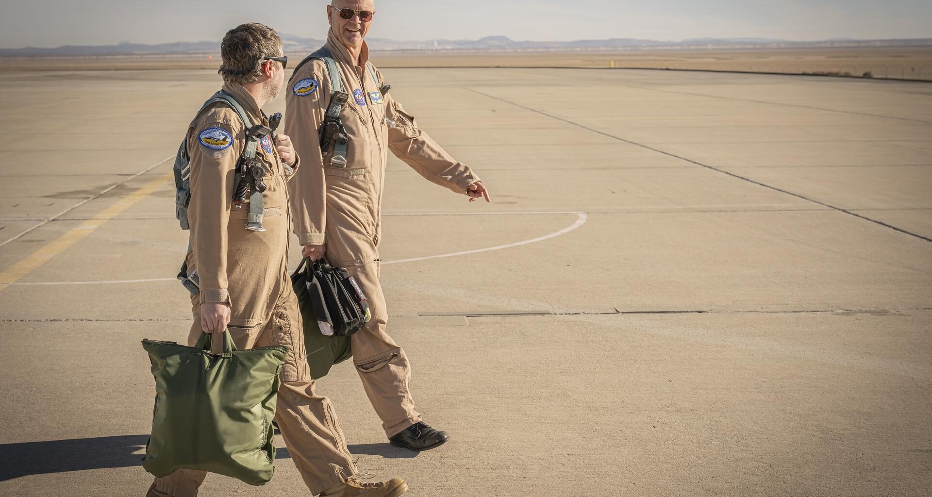 Two men wearing tan flight suits face each other and walk on a concrete surface. The men both carry pilot helmet bags with flight gear inside. Both men are wearing green flight gear.