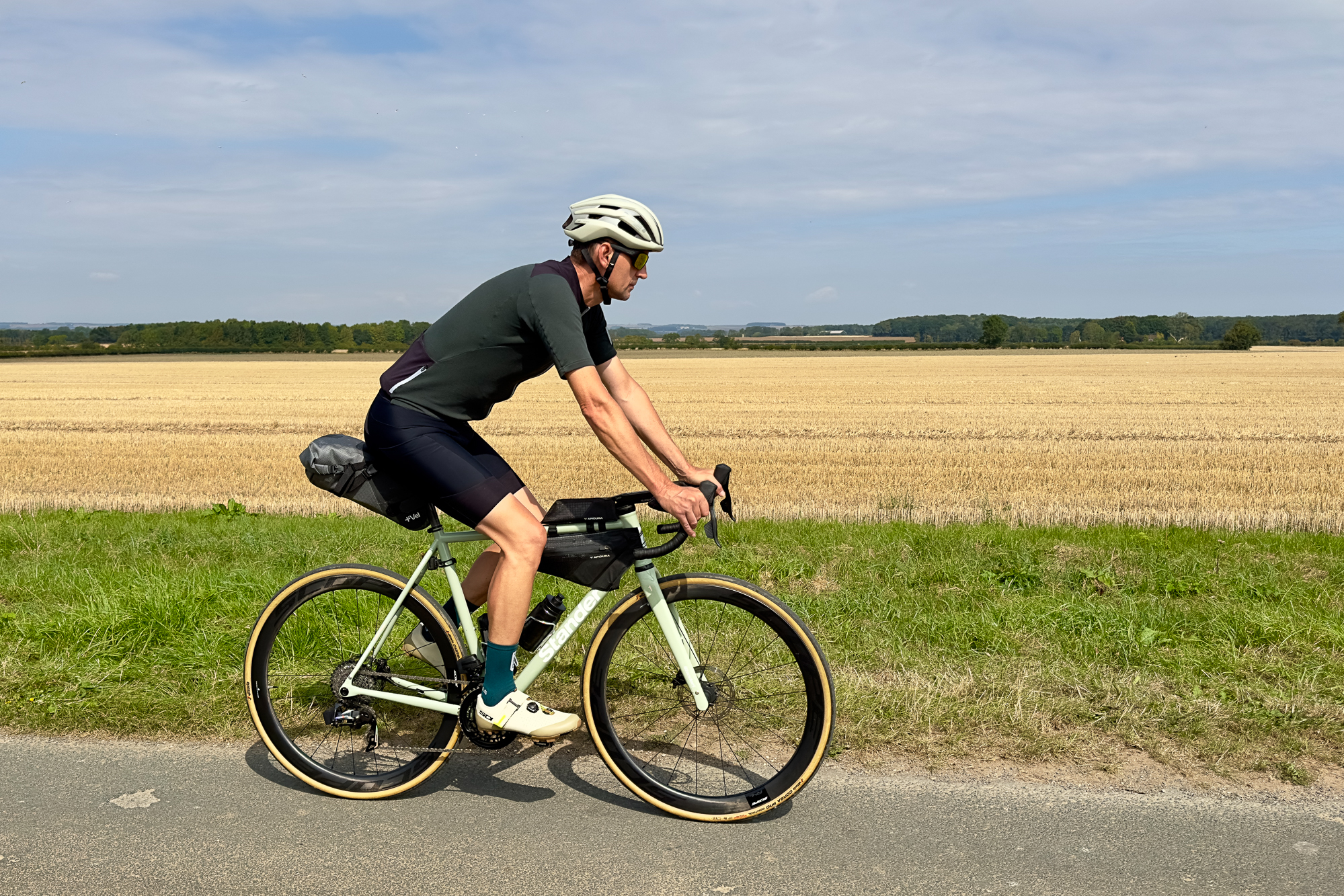 man riding along a quiet rural lane wearing green jersey, beige helmet and black shorts on a pale green bike