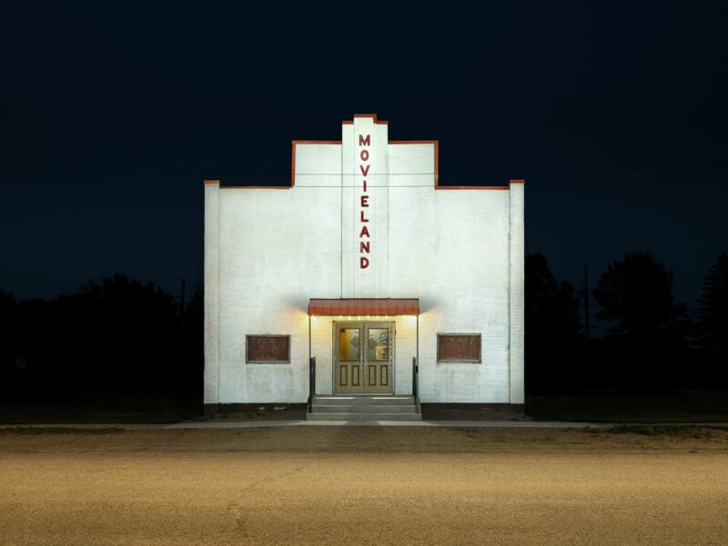 A small, white, rectangular building labeled "MOVIELAND" is brightly lit at night. The building has a central entrance with double doors and a small awning, set against a dark, empty background.