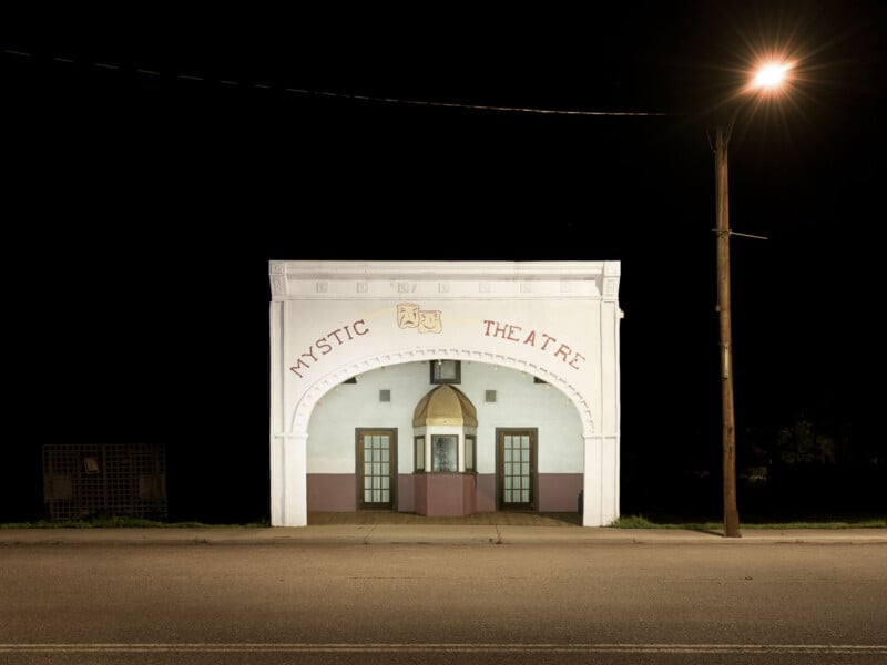 A small, white building labeled “Mystic Theatre” stands alone at night, illuminated by a streetlight. The theater’s arched entrance and ticket booth are visible, with darkness surrounding the building.