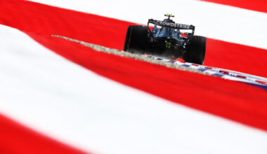 Formula 1 race car speeding along a track, rear view with a CrowdStrike logo on the wing, framed by red and white barrier stripes.