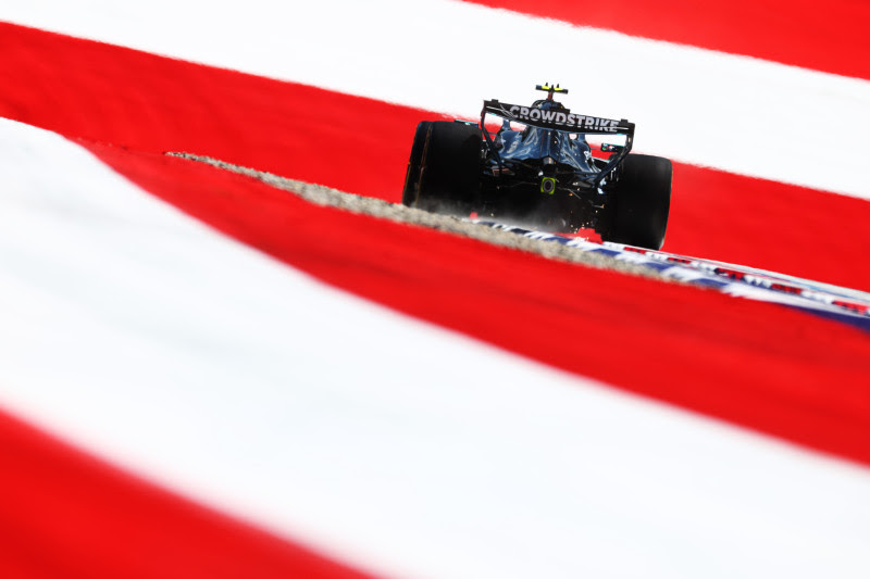 Formula 1 race car speeding along a track, rear view with a CrowdStrike logo on the wing, framed by red and white barrier stripes.
