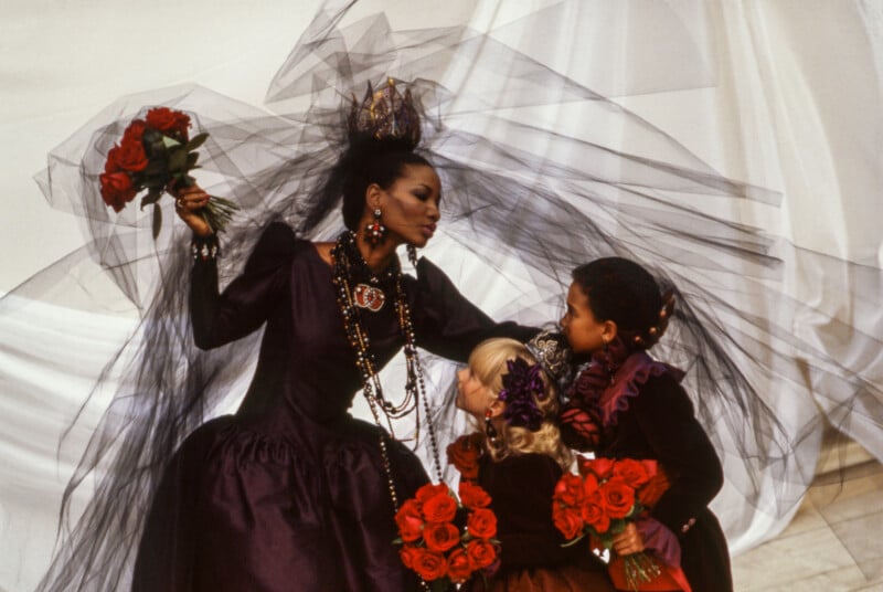 A woman in an elaborate dark gown and black veil holds roses while bending toward two children dressed in matching dark outfits, each holding bouquets of red roses, against a draped white background.