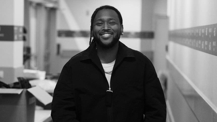 Boi-1da smiles at a camera while pointing towards it in a building alleyway.