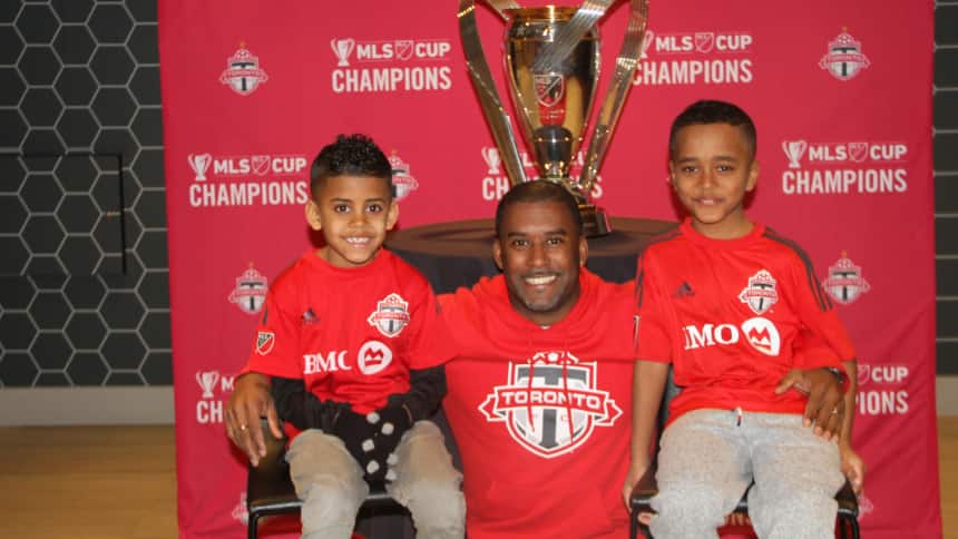 Two children are sitting side by side to their dad in front of a Toronto FC MLS Cup Champions backdrop.