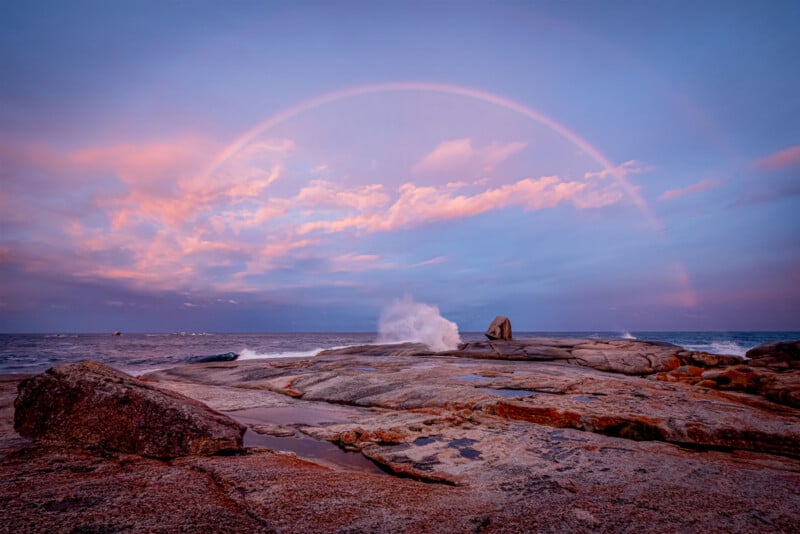 A wide, rocky shoreline with waves splashing against the rocks under a pastel sky. A faint rainbow arcs above, with pink and orange clouds illuminated by the setting or rising sun.