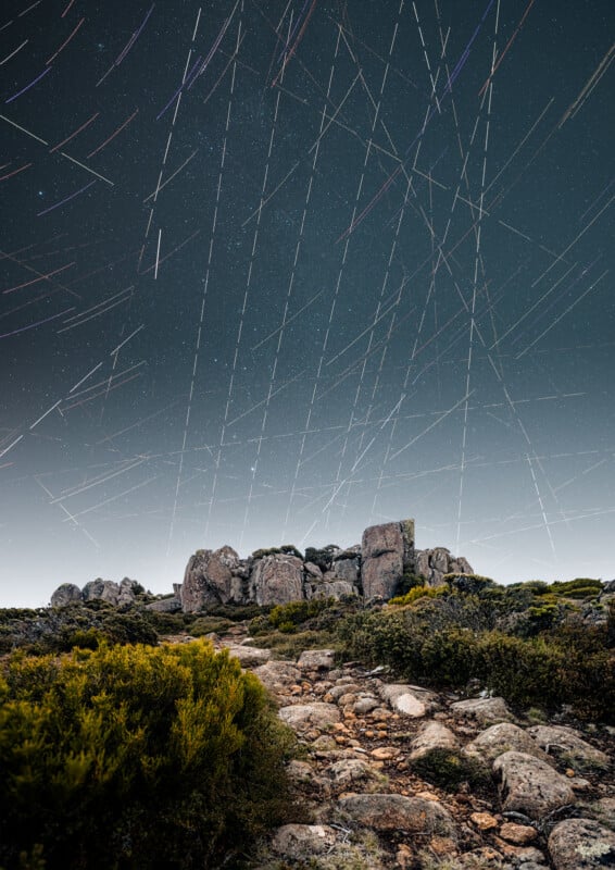 A rocky path leads to large boulders under a night sky filled with colorful star trails, indicating the movement of stars over time. Bushes and greenery line the path in the foreground.