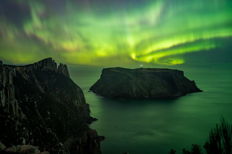 Green and yellow aurora lights illuminate the night sky above a calm sea, with steep rocky cliffs and a flat-topped island visible in the foreground.