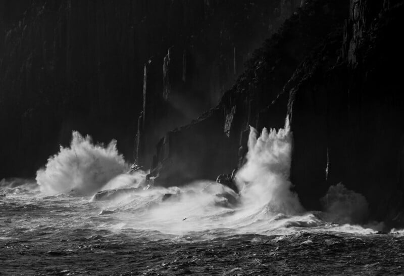 Huge ocean waves crash dramatically against rugged, dark cliffs, sending white spray high into the air. The black and white image emphasizes the contrast and intensity of the natural scene.