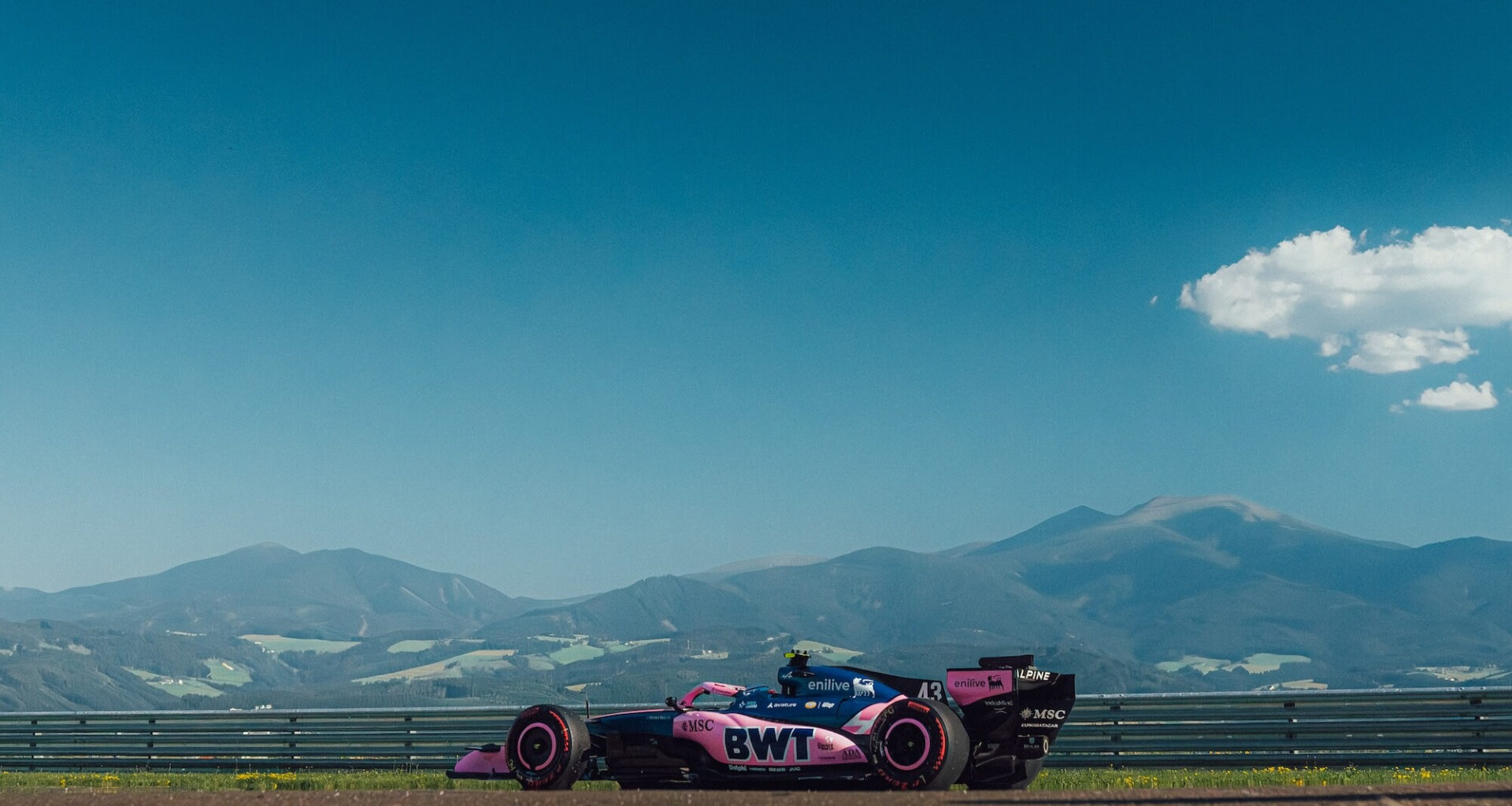 Pink BWT Formula 1 car on a race track with mountains in the distance and a clear blue sky above.