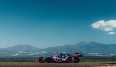 Pink BWT Formula 1 car on a race track with mountains in the distance and a clear blue sky above.